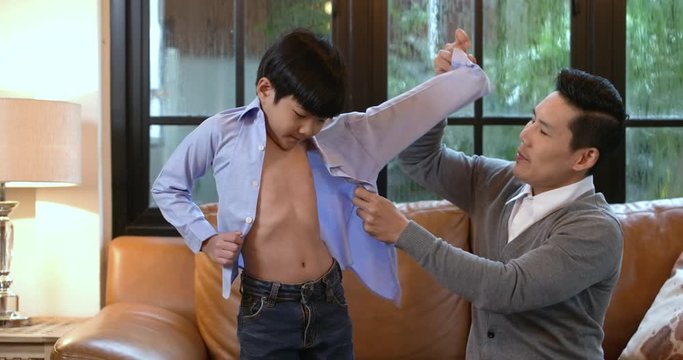 Happy asian father is helping his little son to get dressed, buttoning his shirt in living room.