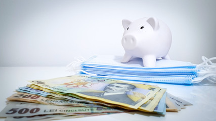 Face masks alongside stack of Romanian money & a white piggy bank

