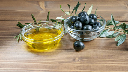 Black olives, olive oil in glass transparent bowls, some olive tree branches on the wooden background in sunlights. Healthy mediterranean food.