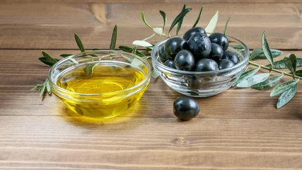 Foto op Plexiglas Black olives, olive oil in glass transparent bowls, some olive tree branches on the wooden background in sunlights. Healthy mediterranean food. © zveruga