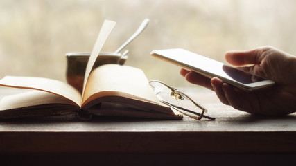 Hand holds smartphone, open old book and cup of coffee near the window