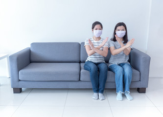 Asian women wearing masks sit together on sofa without space and to cross sign for social distancing to reduce spreading of coronavirus. Work from home, social distancing and covid-19 concept.