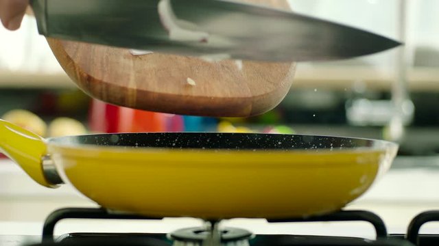 Chopped onions are thrown into the pan with a cutting board for cooking. Yellow pan on the stove, oil in the pan splashing. Slow motion.