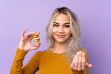 Teenager girl over isolated purple background holding colorful French macarons and inviting to come