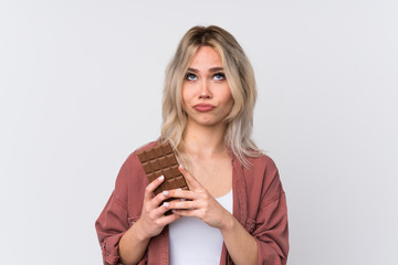 Teenager blonde girl over isolated white background taking a chocolate tablet and having doubts