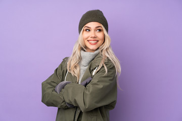 Teenager blonde girl with winter hat over isolated purple background with arms crossed and happy