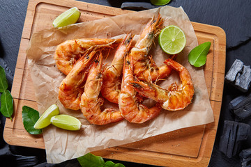 Top view of grilled shrimps with lime on wooden cutting board on black stone tabletop. 