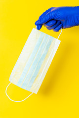 A hand in a blue latex glove holds a surgical mask on a yellow background.