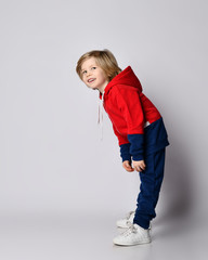 Happy smiling blond kid boy in blue and red hoodie and pants poses sideways bending over reaching his hands to toes