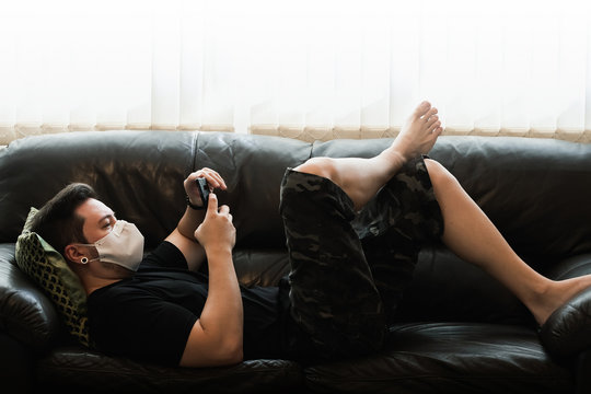 Man wearing a face mask in quarantine at home using the phone lying on sofa. Man on quarantine browsing the mobile internet at home.