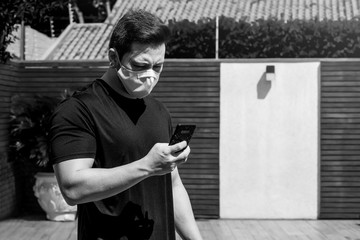 Standing man wearing an white face mask during quarantine, at the home yard, checking the news on the phone. 