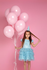 adorable smiling little child girl in blue dress and birthday hat celebrating with pastel pink air balloons on pink background. birthday party.