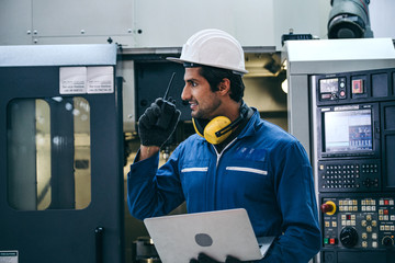 Male industrial engineer or technician in safety hard helmet and uniform using laptop checking on machine and radio phone calling team worker, heavy technology invention industry manufacturing factory