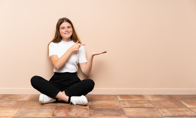 Ukrainian teenager girl sitting on the floor holding copyspace imaginary on the palm to insert an ad