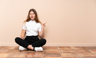 Ukrainian teenager girl sitting on the floor surprised and pointing side