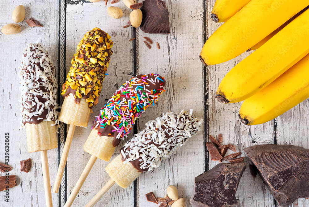 Poster frozen chocolate dipped banana pops with coconut, nuts and sprinkles. above view table scene over a 