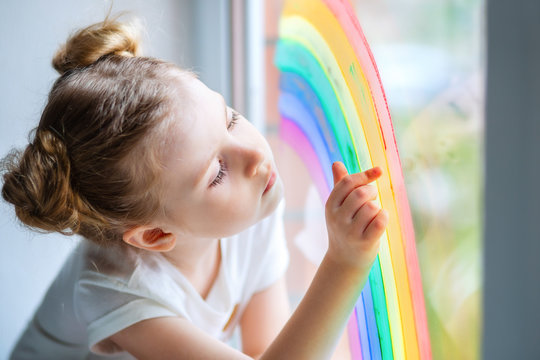 A little girl with blond hair looks at a rainbow on the window. 