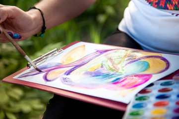 close-up of a man holding paint and a brush, drawing a picture of bright watercolor paints