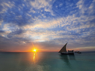 Calm indian ocean in the rays of the setting sun and blue sky with small clouds. a boat with a sail goes on the ocean.