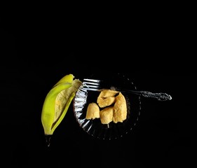 Plate with banana pieces and fork on black background. diet food