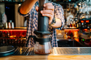 Fototapeta na wymiar Alternative Coffee Brewing Method. Close-up of the hands of barista who is preparing filter coffee in an aeropress. Barista wears a checkered flannel shirt and works at the bar counter.