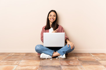 Young mixed race woman with a laptop sitting on the floor pointing to the side to present a product