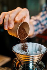 Vertical image. Alternative Coffee Brewing Method. Close-up of the hands of barista, pouring ground coffee from a small cup into a coffee filter