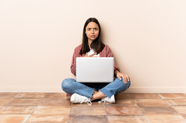 Young mixed race woman with a laptop sitting on the floor pointing to oneself