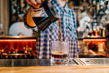 Alternative Coffee Brewing Method. 
Close-up of the hands of barista, pouring a black filter coffee from a teapot into a transparent glass on a bar counter in a cafe.