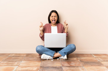 Young mixed race woman with a laptop sitting on the floor with fingers crossing