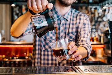 Alternative Coffee Brewing Method. 
Close-up of the hands of barista, pouring a black filter coffee from a teapot into a transparent glass on a bar counter in a cafe.
