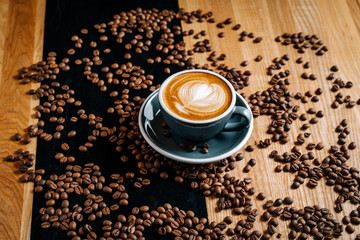 Gray cup of coffee on the black and brown table among spilled roasted coffee beans. Cappuccino with cofee art in the cafe.