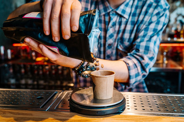 Close-up of the hands of a barista, wearing flannel shirt, who sprinkles coffee beans from a black packet into a small brown cup, standing on the scales on a bar counter