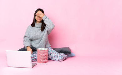 Young mixed race woman eating popcorn while watching a movie on the laptop covering eyes by hands. Do not want to see something