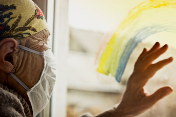 Grandmother is self-insulating while looking at the street through the window pane. An elderly woman in a mask looks at the street through a colored rainbow on the glass. Stay home - be safe. 