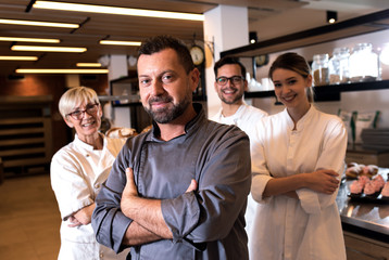 Portrait of mid age male baker at bakery with his colleagues in backgrounds.