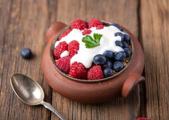 Granola with white yogurt with raspberries and blueberries in ceramic bowl on natural wooden desk.