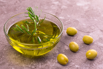 Olive oil in a transparent bowl and a branch of rosemary on a gray background.