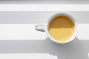 White espresso coffee mug. Light effect, white and dark stripes fall on the subject and background. Light and shadow in the composition. A cup of coffee near the window.