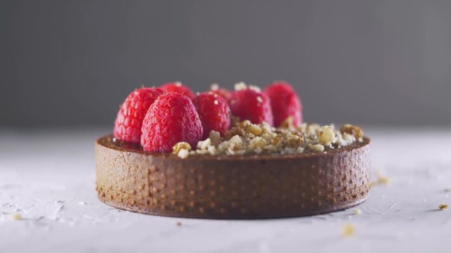 Chipped nuts falling down to the appetizing chocolate tart with raspberries and nuts on the grey background, real time