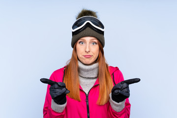 Skier redhead woman with snowboarding glasses over isolated blue wall pointing to the laterals...