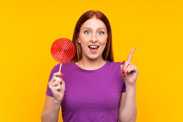 Young redhead woman holding a lollipop over yellow background pointing up a great idea