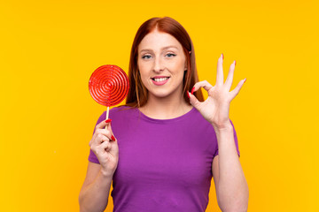 Young redhead woman holding a lollipop over yellow background showing ok sign with fingers