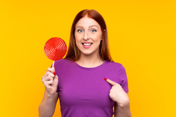 Young redhead woman holding a lollipop over yellow background with surprise facial expression