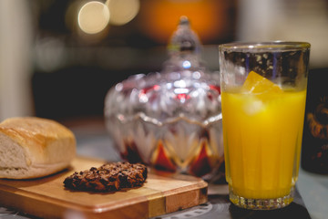 Glass of orange juice and ice, wood table with bread and handmade cookie, and glass bonbonniere with warm light