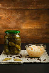 Jars with pickled cucumbers and sauerkraut on a wooden table on a black background.