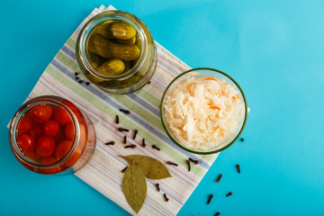 Jars with pickled cucumbers and tomatoes and sauerkraut on a wooden table on a blue background on a linen towel.