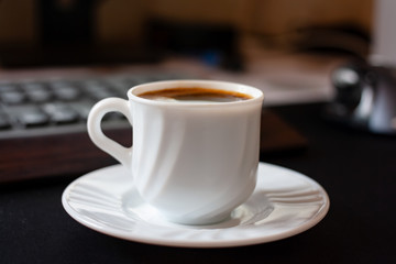white espresso cup on a saucer in front of a keyboard in the workplace. Hot coffee while working day. Cozy calm atmosphere at the workplace. Fragrant refreshing drink in mug. Coffee break concept