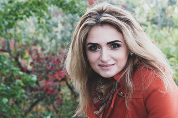 Naklejka premium portrait of attractive smiling woman with blond long hair in orange coat looking at camera in front of bushes with green and red leaves in forest