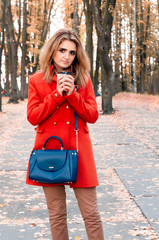 attractive woman in orange coat with blue bag and long hair holding paper cup and looking at camera  in front of trees with yellow foliage in park